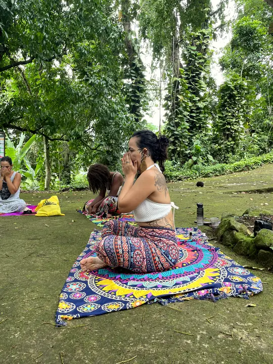 Mulher sentada sobre uma canga colorida, de olhos fechados, em uma prática de respiração ao ar livre