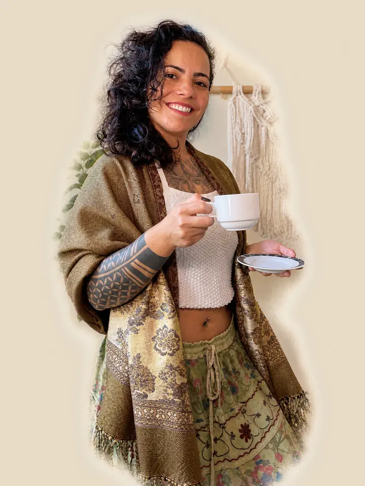 Mulher sorrindo com uma xícara de café, usando uma pashmina estampada, em frente a um macramê