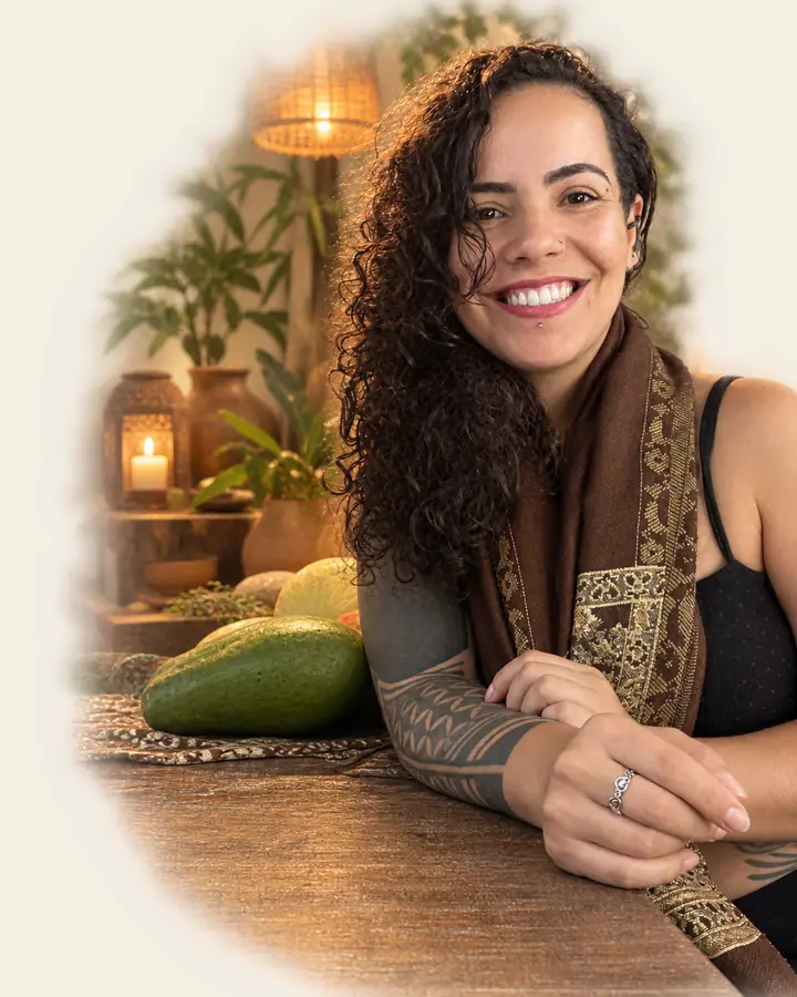 Mulher sorrindo, apoiada em uma mesa de madeira com frutas, em um ambiente aconchegante com plantas e velas